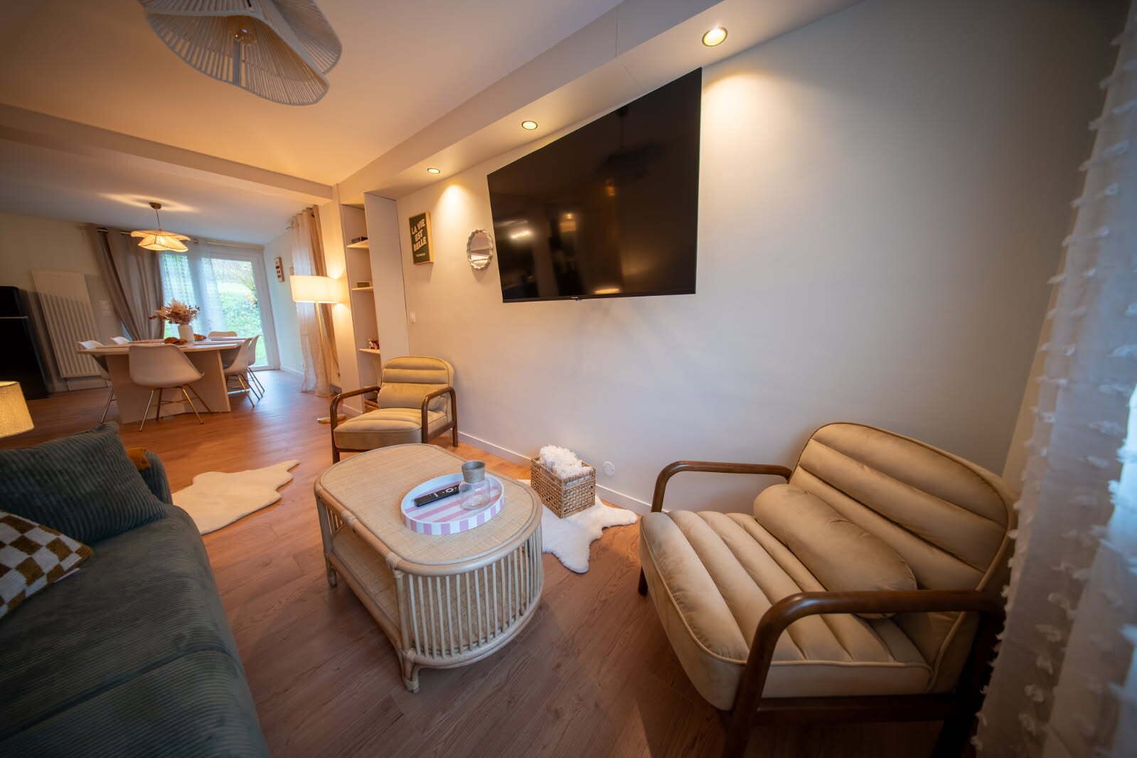 Un salon moderne comprenant un canapé, un fauteuil et un pouf, un coin repas avec une table et des chaises, et un téléviseur à écran plat fixé au mur.