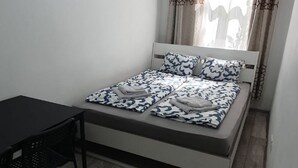 Een eenpersoonsbed met beddengoed met een patroon, een klein tafeltje en een stoel in een kamer met een raam en gordijnen.