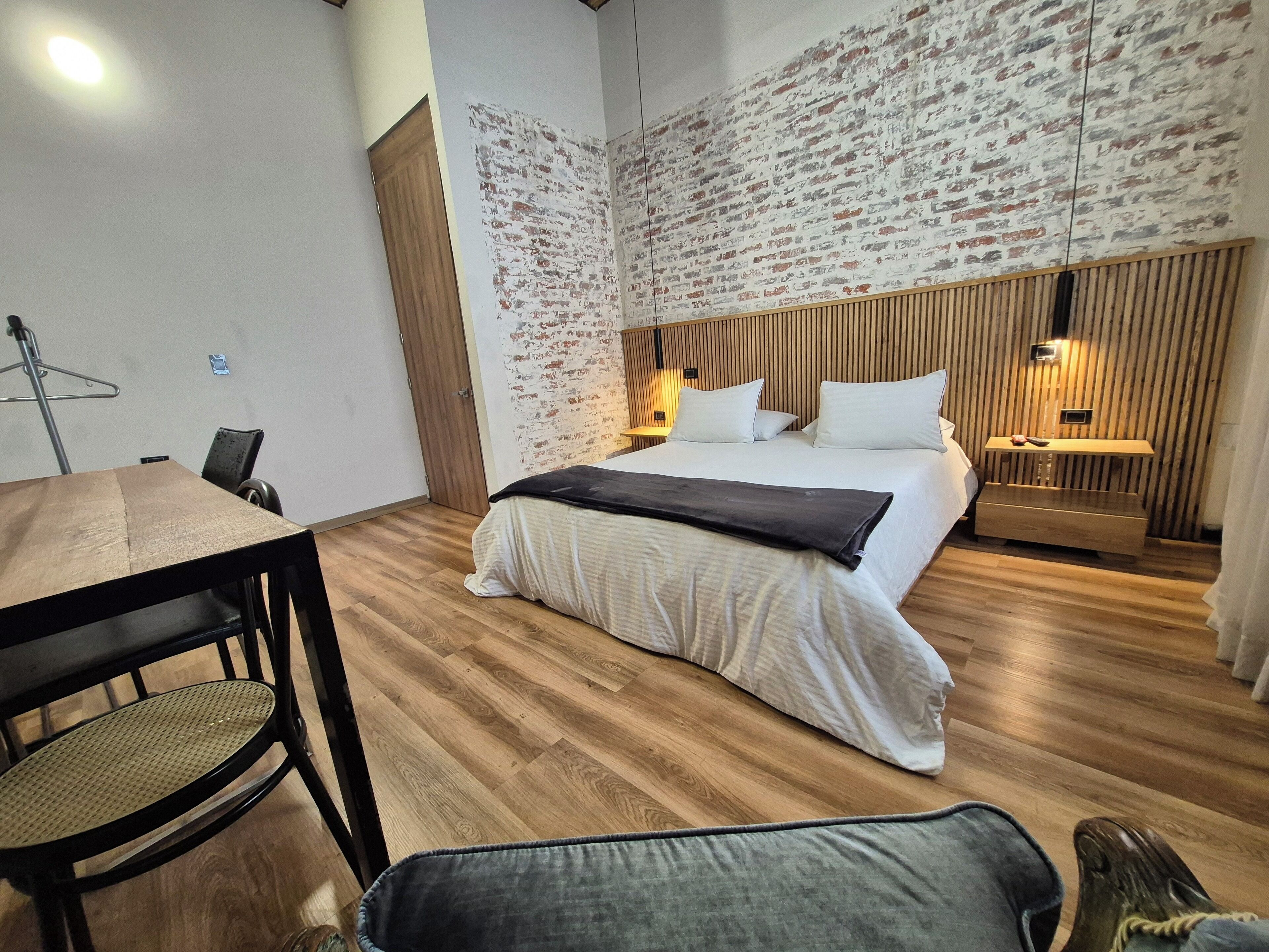 Un dormitorio moderno con suelo de madera, una cama con ropa blanca, un cabecero de madera y una mesita de noche con lámpara.