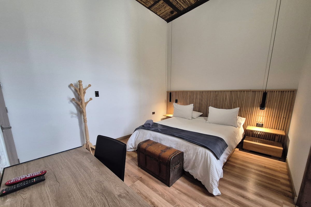 A modern bedroom with a wooden headboard, a bed with white linens, a bedside table with a lamp, a wooden coat rack, and a small wooden chest.