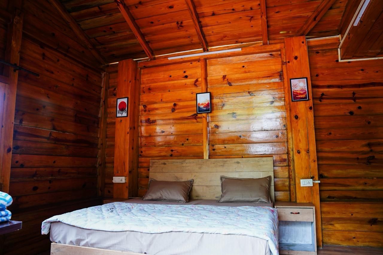 A wooden cabin bedroom with a bed, two pillows, a nightstand, and a wall-mounted shelf with pictures.