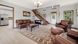 A spacious living room with a brown leather sofa, a coffee table, a patterned rug, a chandelier, and a staircase leading to another level.