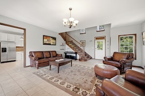 A spacious living room with a brown leather sofa, a coffee table, a patterned rug, a chandelier, and a staircase leading to another level.