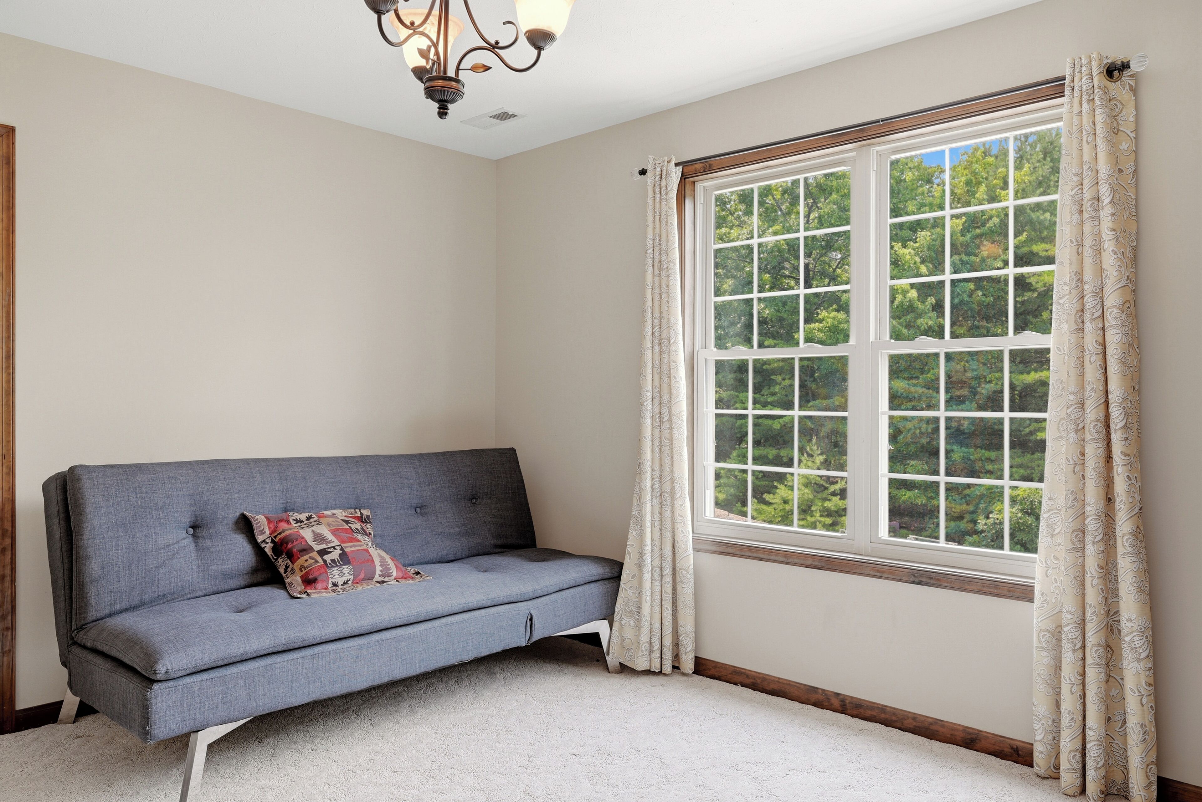 A room with a grey sofa, a patterned cushion, a large window with curtains, and a chandelier.