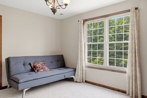 A room with a grey sofa, a patterned cushion, a large window with curtains, and a chandelier.