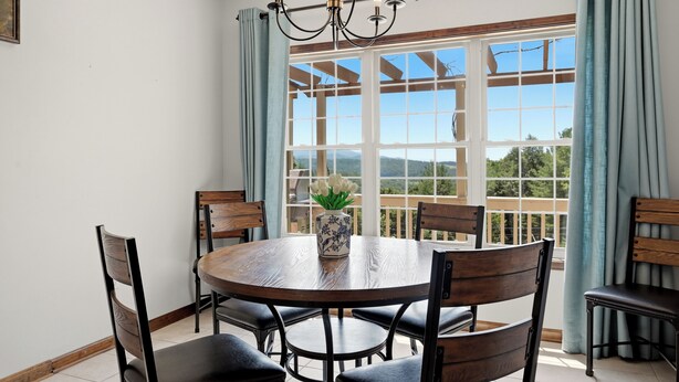 A dining area with a round table, chairs, a chandelier, and a view through large windows.