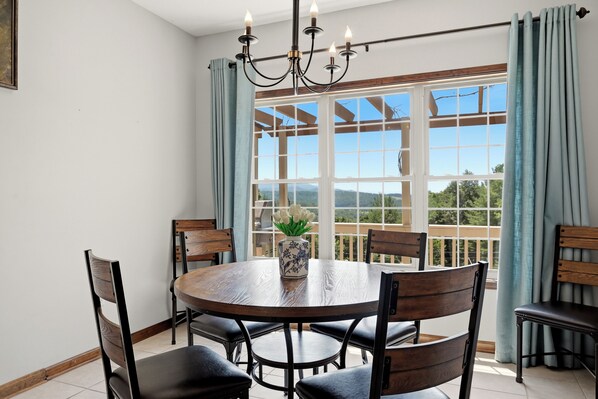 A dining area with a round table, chairs, a chandelier, and a view through large windows.