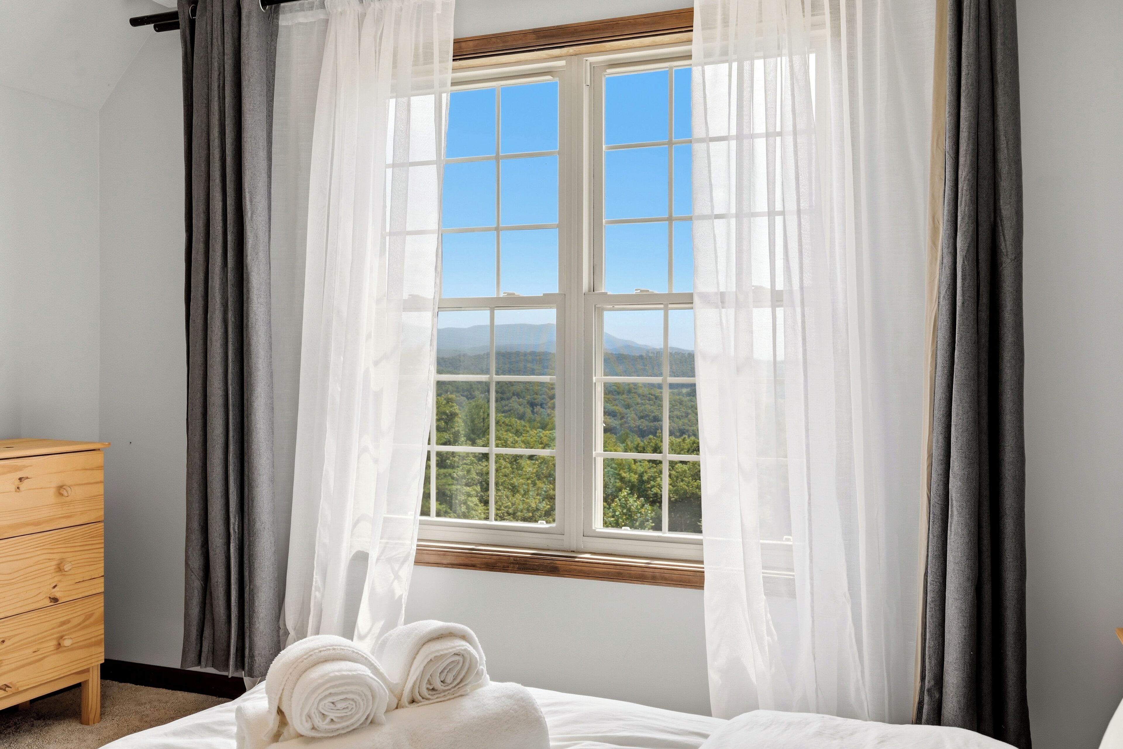 A bedroom with a large window, a wooden dresser, and two rolled towels on the bed.