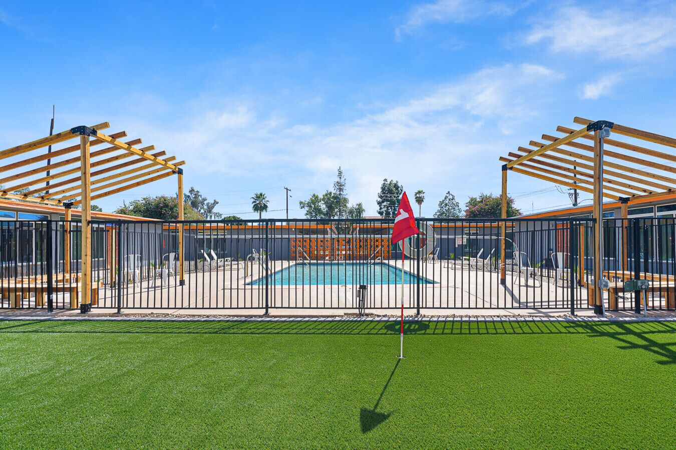 Een omheind zwembadgedeelte met een vlag, een houten pergola en kunstgras.