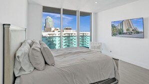 A modern bedroom with a large bed, a view of a cityscape through floor-to-ceiling windows, and a framed picture on the wall. - Oceanview Penthouse Pool Beach Access (Hollywood)