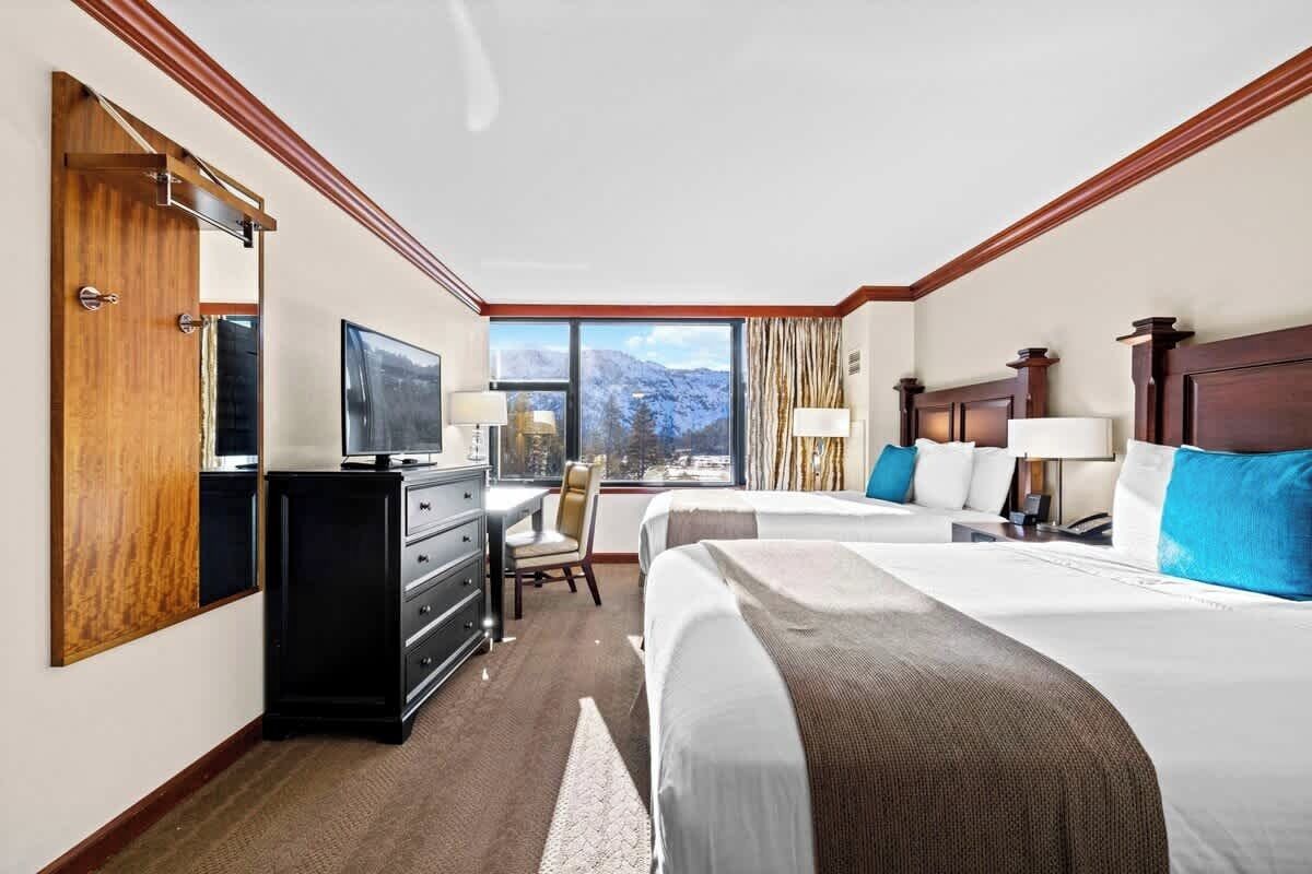 A hotel room with two beds, a desk, a TV, and a window with a mountain view.