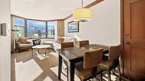 Una habitación de hotel moderna con zona de comedor, un conjunto de sofás, un ventanal con vistas a la montaña y una puerta de madera.