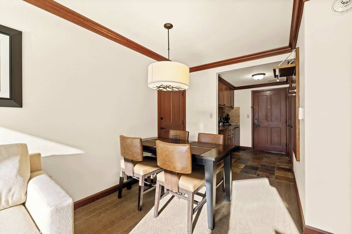 A dining area with a wooden table and chairs, a hanging light fixture, and a kitchen visible through an open door.