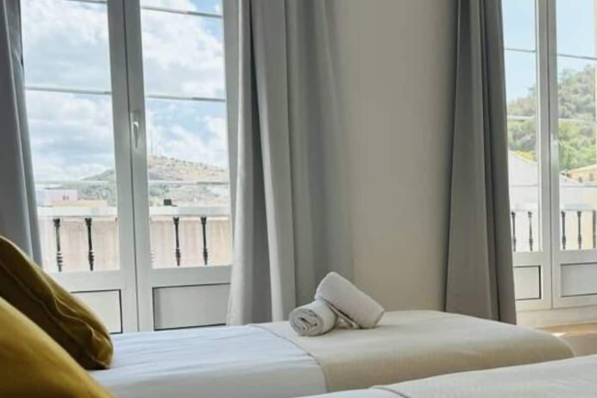 Habitación de hotel con dos camas, vistas a la ciudad a través de grandes ventanales y un balcón con barandilla.