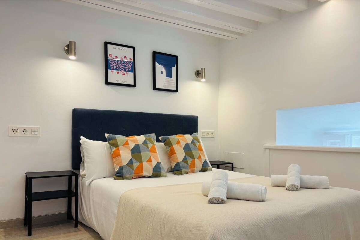 Habitación de hotel con cama, dos mesitas de noche, cabecera y dos cuadros en la pared.