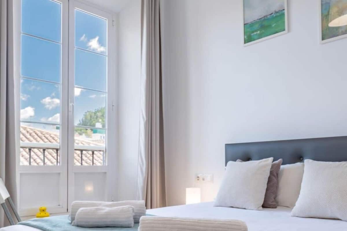 Habitación de hotel con cama, ropa de cama blanca, manta azul y vista al cielo y edificios a través de una ventana.