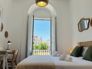 Habitación de hotel con cama, escritorio, silla, balcón y vistas a edificios, además de una lámpara en el techo.