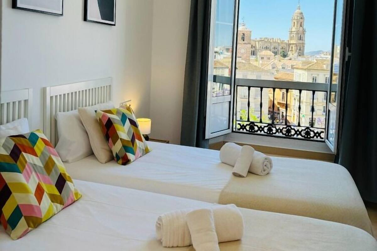 Habitación de hotel con dos camas, un ventanal con vistas a un paisaje urbano y arte abstracto en la pared.