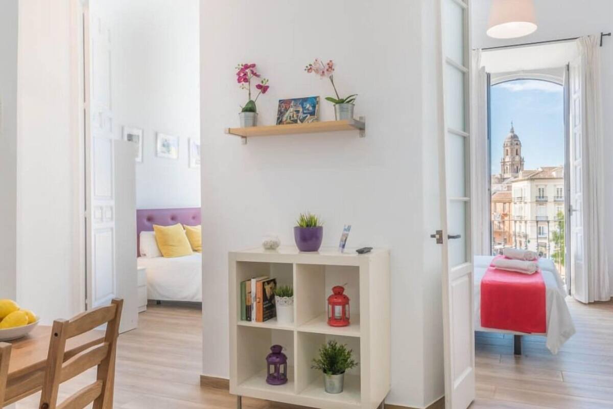 Una habitación de hotel moderna con suelo de madera, una cama blanca con manta roja, una mesita con un cuenco de limones, una estantería blanca con adornos y vistas a edificios a través de un ventanal abierto.