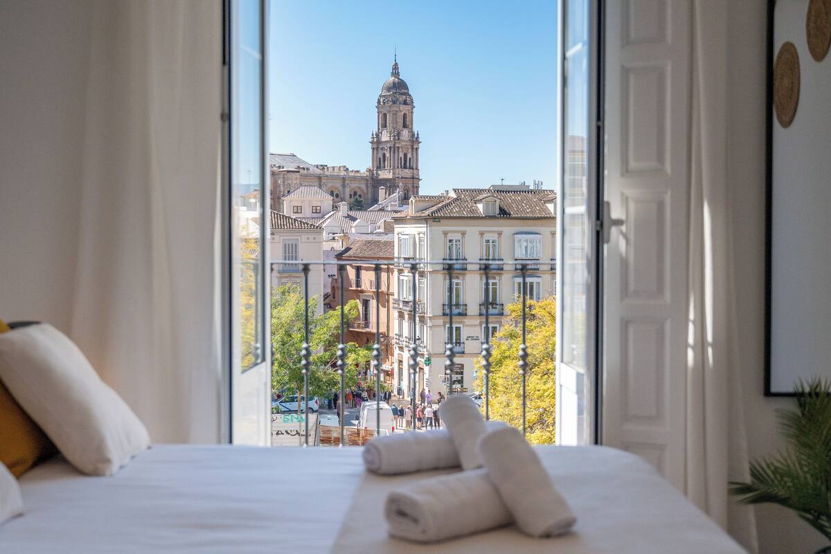 Habitación de hotel con cama, almohadillas, vista a la ciudad con una torre destacada y balcón con barandillas.