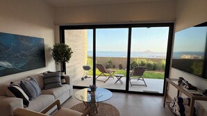 Uma sala de estar moderna com sofá, poltrona e mesa de centro de vidro. Há uma televisão grande em um suporte, e uma varanda com vista para o mar e edifícios.