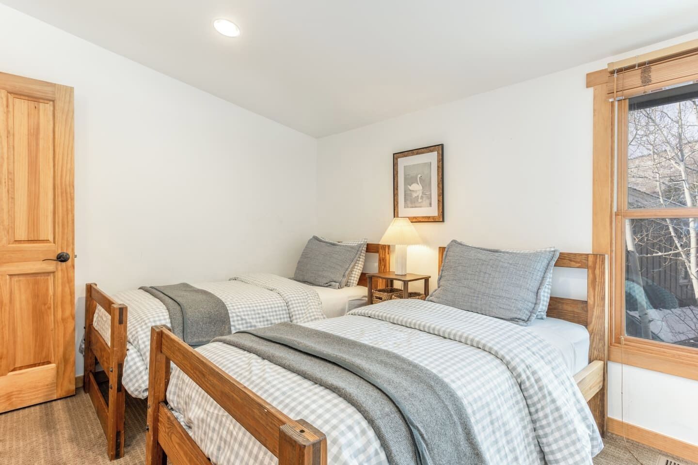 Une chambre à coucher avec deux lits en bois, une table de chevet avec une lampe, un tableau encadré au mur et une fenêtre donnant sur des arbres.