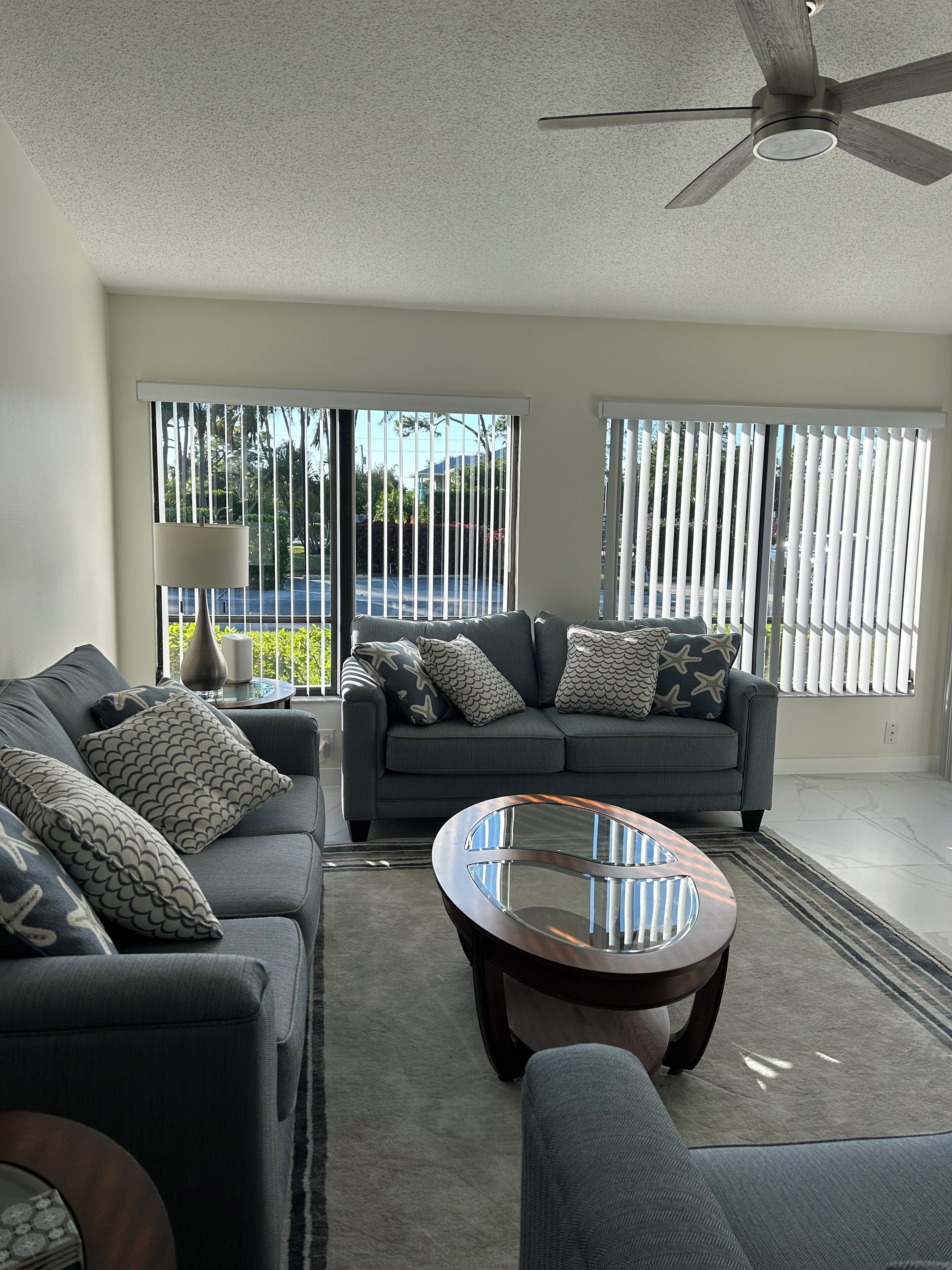 A modern living room with a grey sofa, patterned cushions, a glass coffee table, and a ceiling fan.