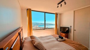 A bedroom with a bed, a television, and a window with a view of a city and sea. - Ocean Front House 84 (Valparaíso)