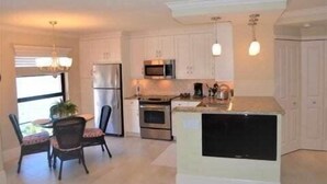 A compact kitchen with white cabinets, a refrigerator, a microwave, an oven, and a sink. There is a dining area with a table and chairs. - Great views of Marco River from 5th Floor condo (Marco Island)