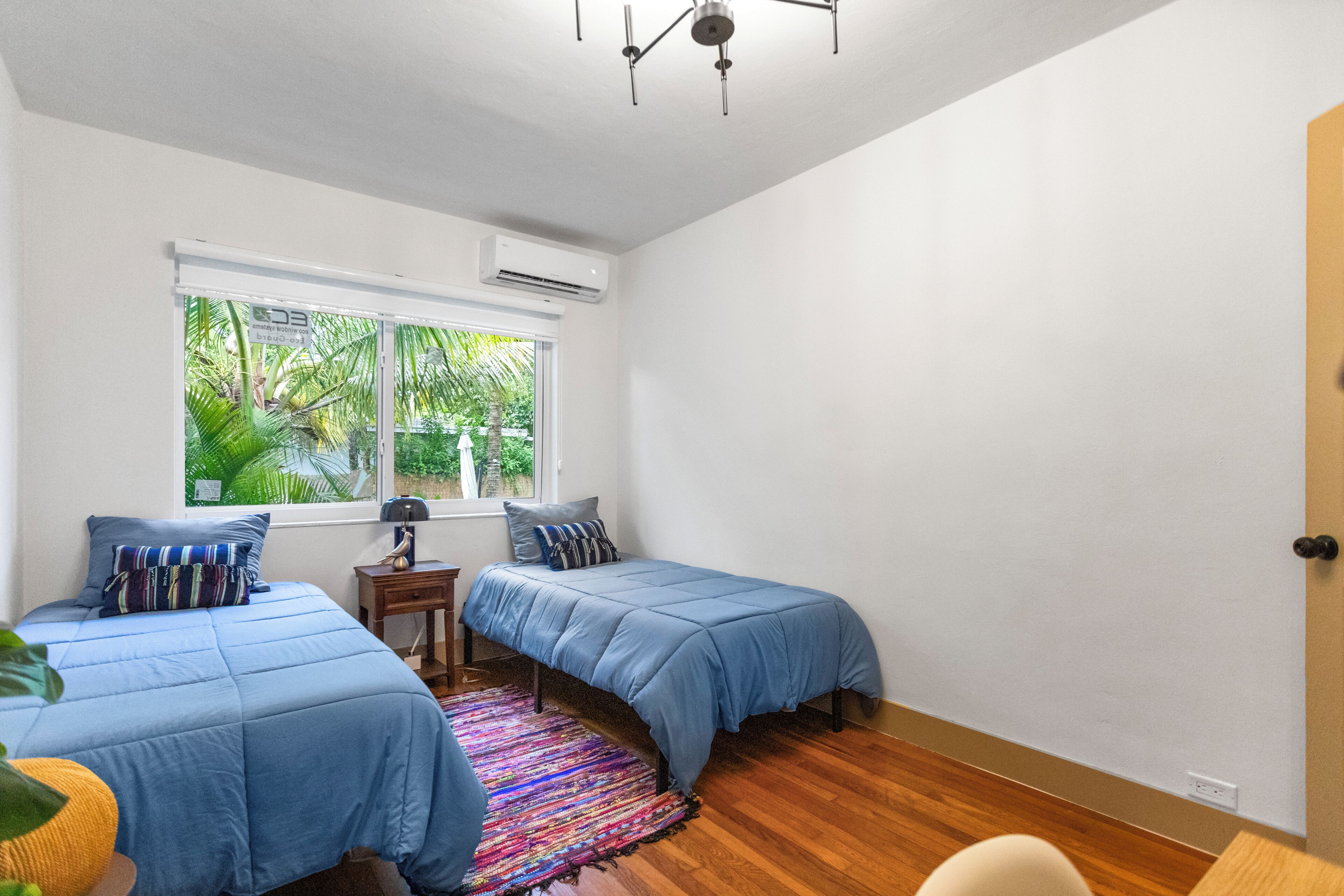 A bedroom with two beds, a window with a view of greenery, a small table with a lamp, and a ceiling fan.