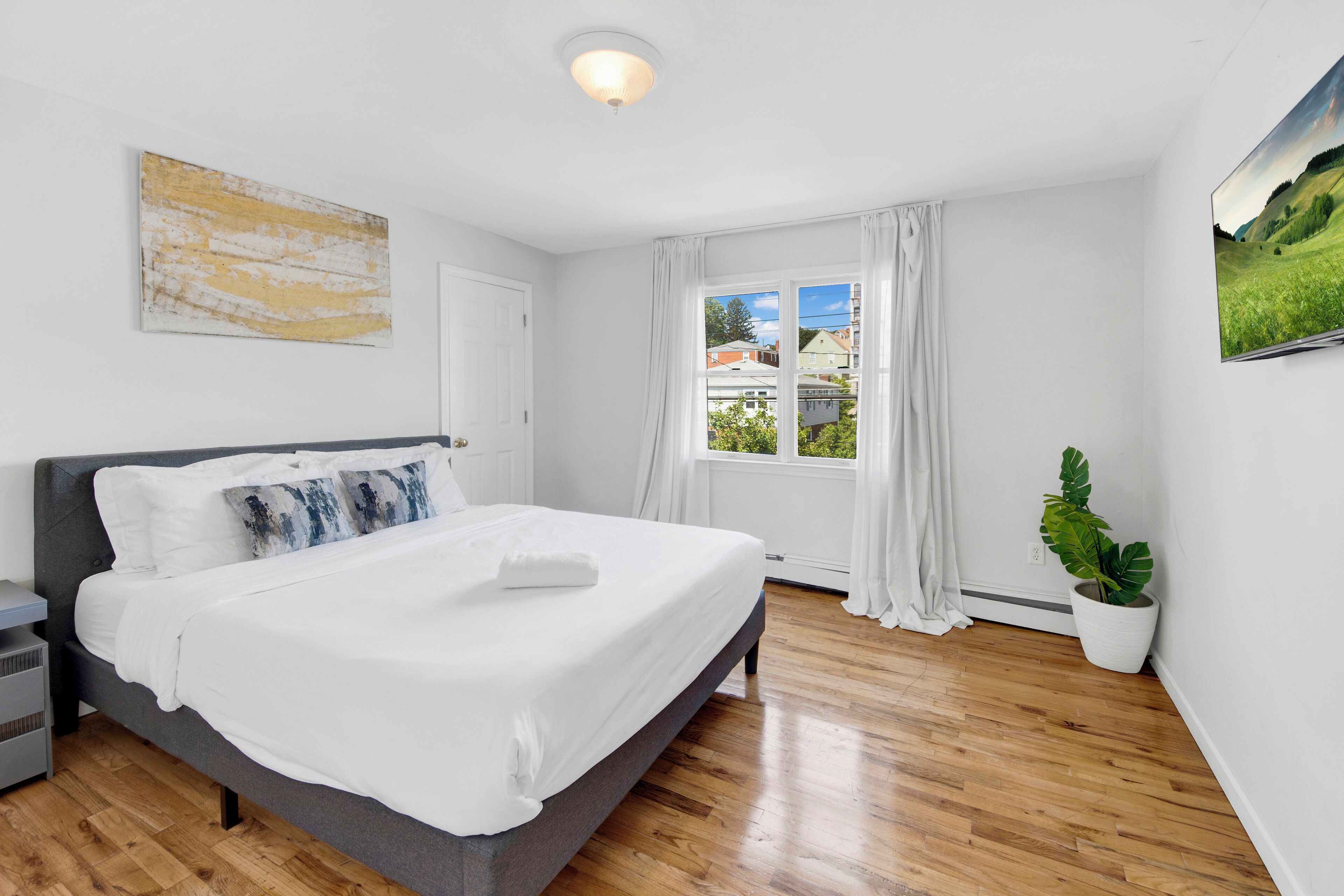 Une chambre moderne avec un grand lit, une table de chevet, une plante en pot et une fenêtre avec des rideaux.