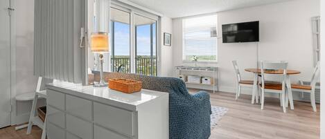 A modern living room with a white sofa, a dining table with chairs, a TV mounted on the wall, and a balcony with a view.
