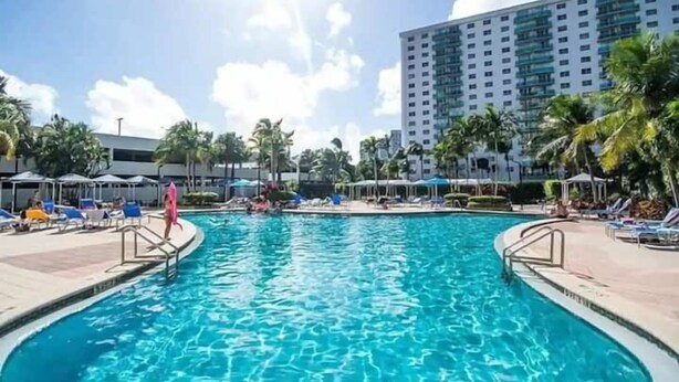 A swimming pool with lounge chairs and umbrellas, surrounded by palm trees and a tall building in the background.