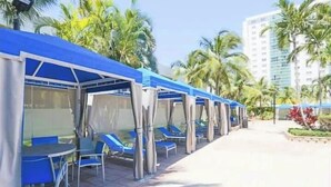 A covered outdoor seating area with blue chairs and tables, surrounded by palm trees and a building in the background.