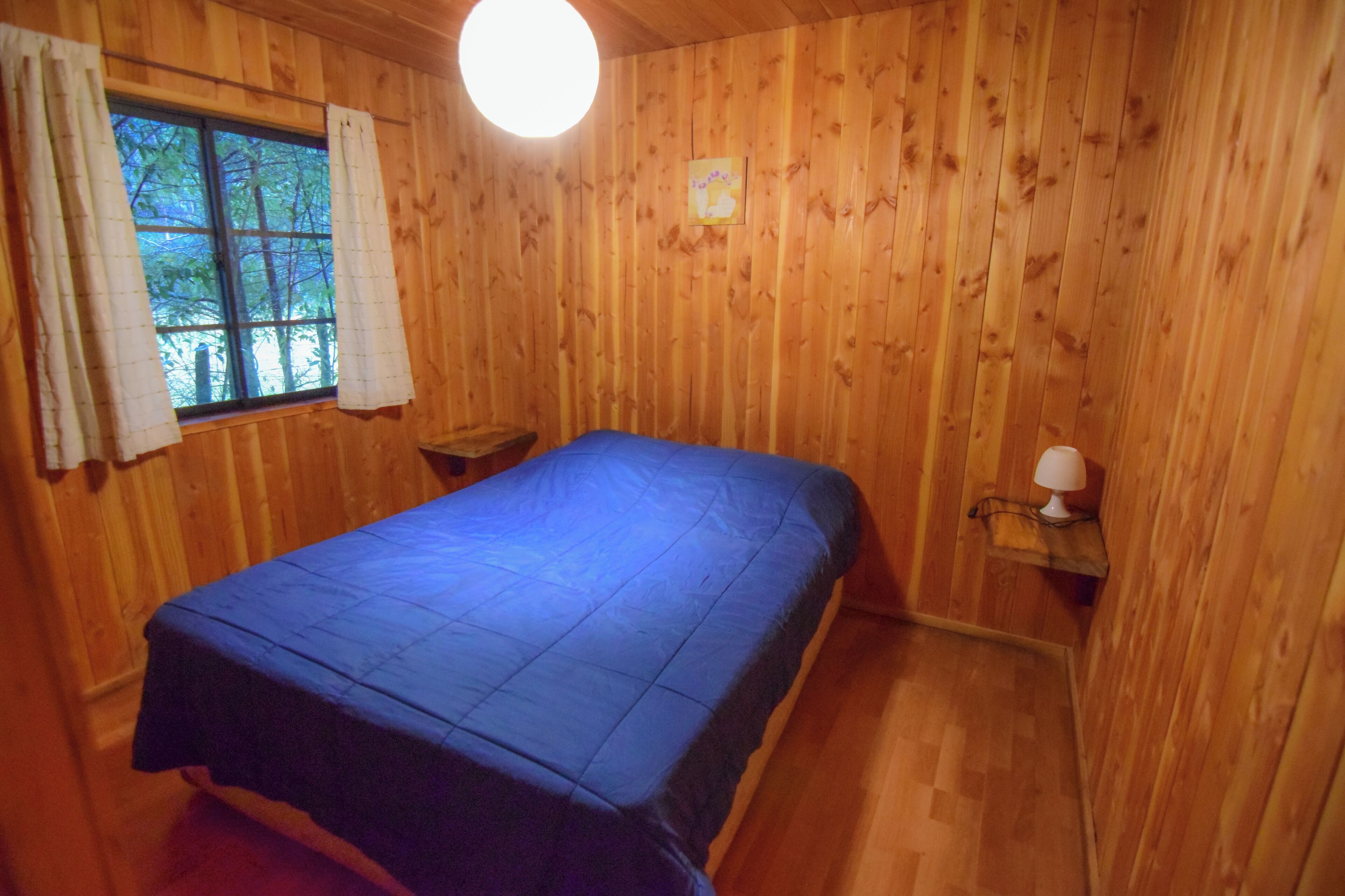 Une pièce aux murs lambrés, avec un lit, une fenêtre avec des rideaux et une lampe fixée au mur.