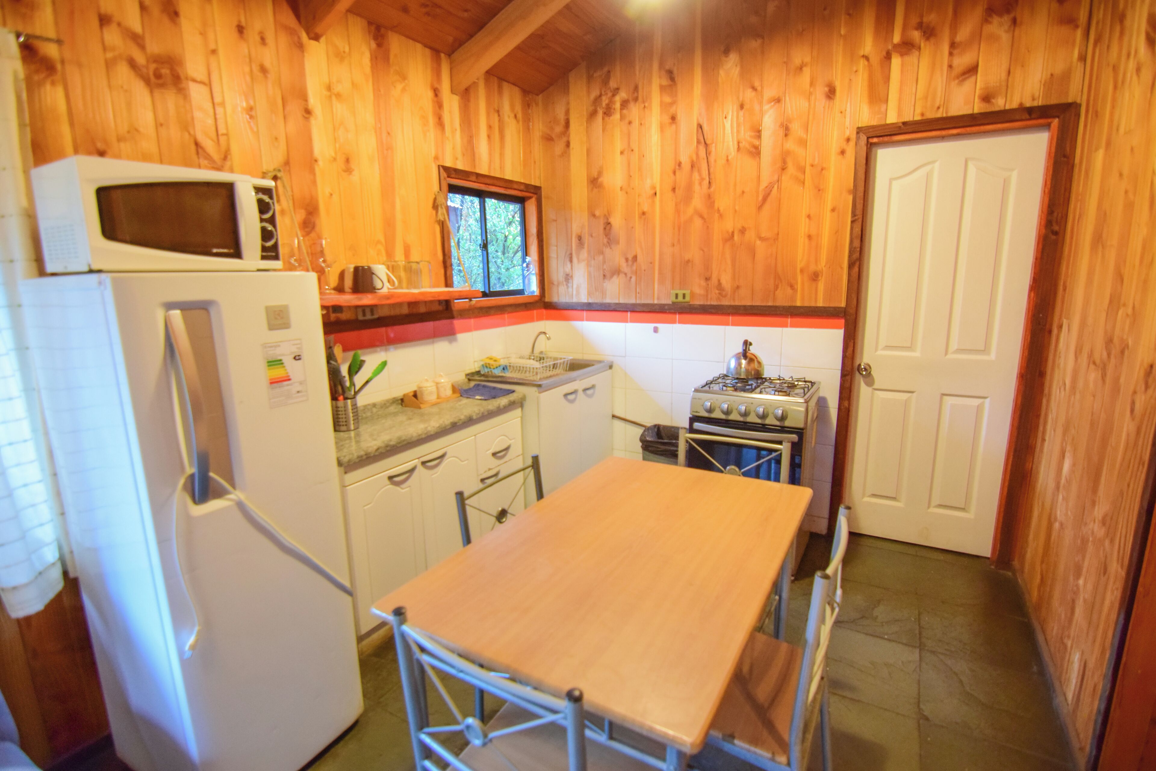 A compact kitchen with wooden walls, a white refrigerator, a microwave, a sink, a stove, and a wooden table.
