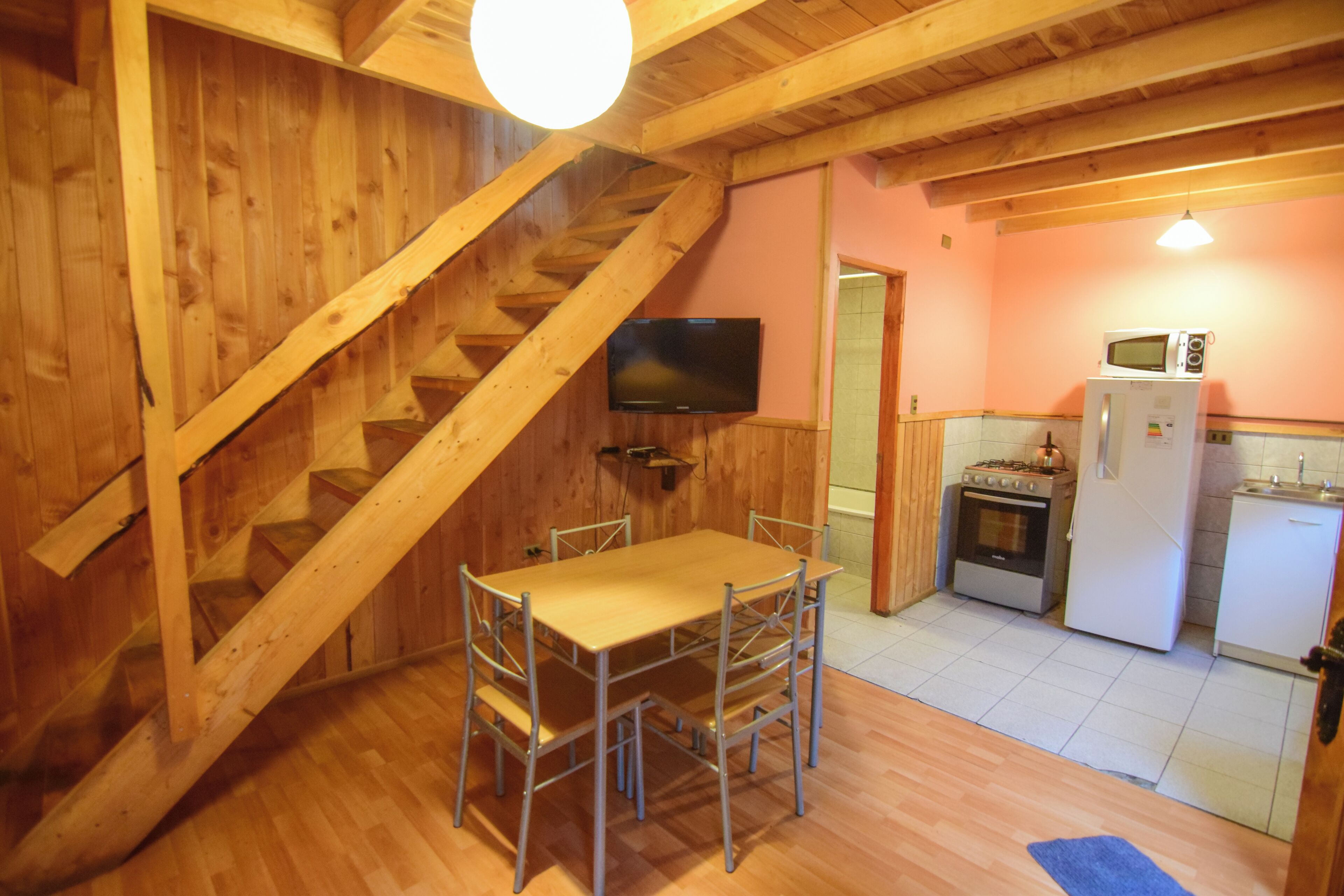 A wooden interior with a dining table and chairs, a kitchen area with appliances, and a staircase.