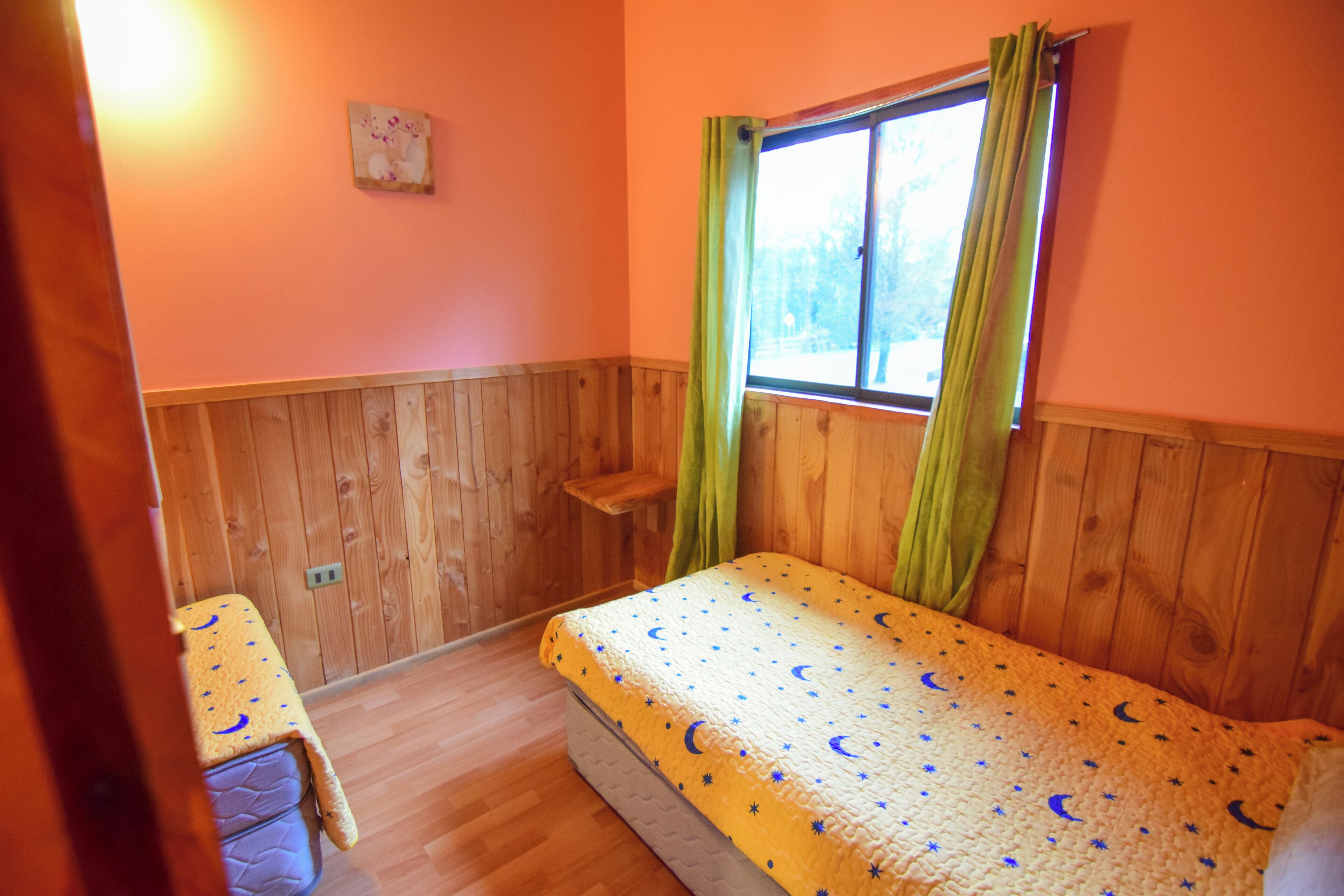 Una habitación con paredes de madera, una cama con cubrecama estampado con estrellas, una ventana con cortinas verdes y una mesita con mantel amarillo.