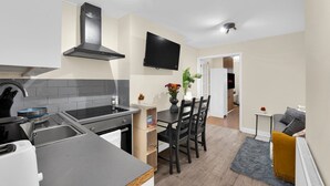 A modern kitchen with a stove, sink, and black range hood. There is a dining table with chairs and a small plant on it. A flat-screen TV is mounted on the wall above the table. - Peaceful Kent Stay with Garden & Parking (Dartford)