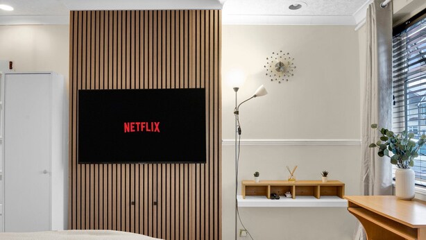 A modern bedroom with a mounted TV displaying 'NETFLIX', a bedside lamp, a small shelf with decorative items, and a window with blinds. - Peaceful Kent Stay with Garden & Parking (Dartford)