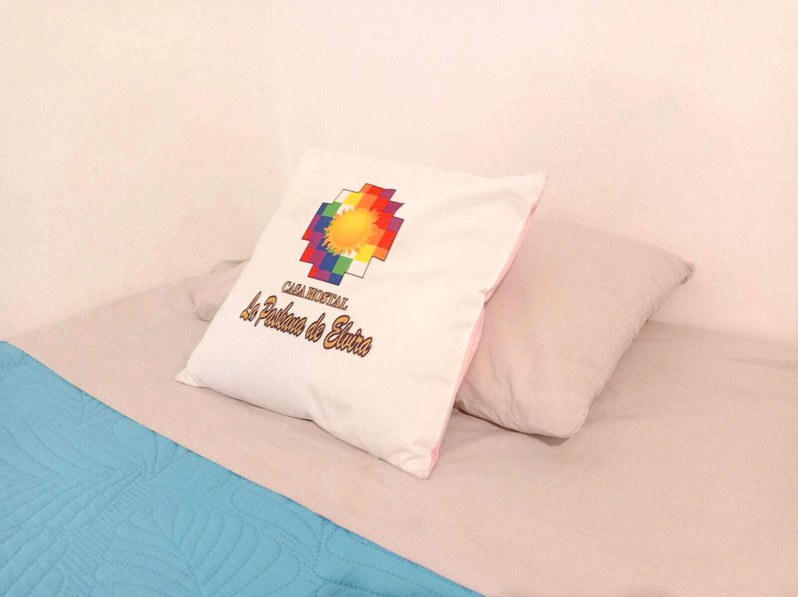 A pillow with a colorful design and text on a bed with a blue quilt.