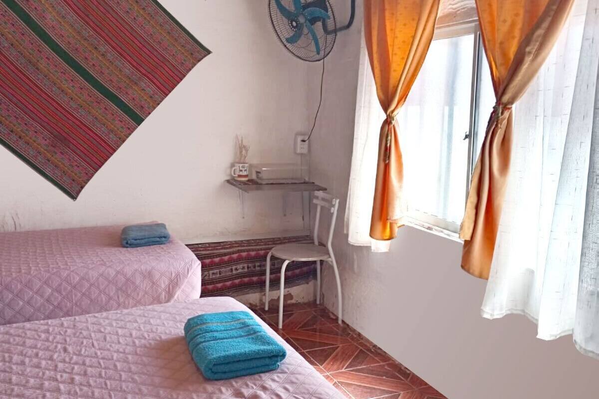 Ein Zimmer mit zwei Betten, einem Ventilator, einem Schreibtisch und einem Fenster mit Vorhängen.