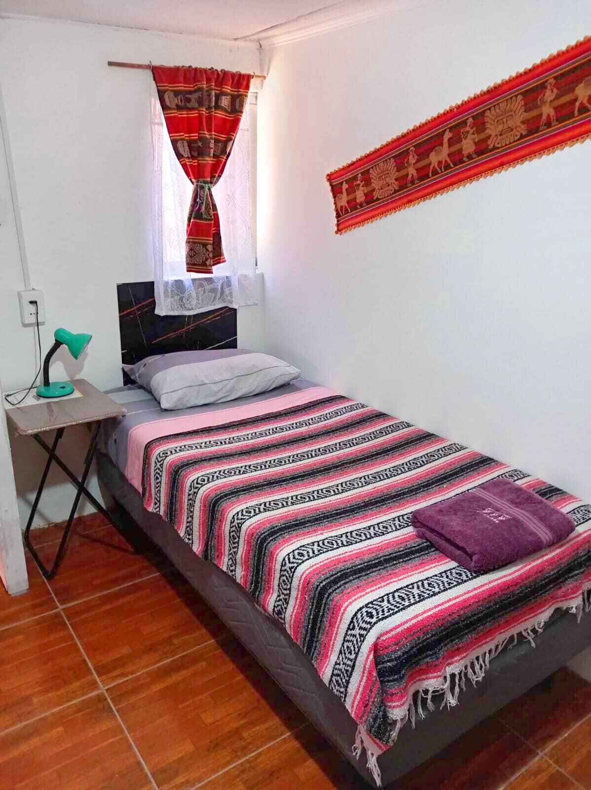 A small, neatly arranged bedroom with a bed, a desk, a lamp, and a window with curtains.
