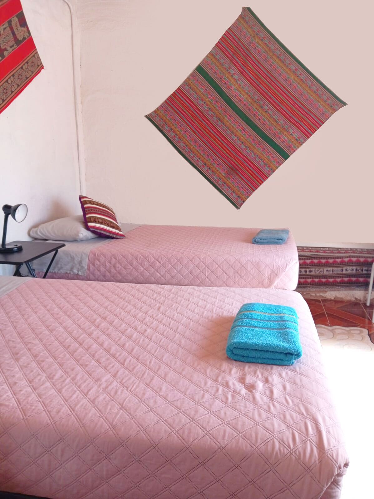 Two beds with pink quilts, a red and green patterned wall hanging, a white pillow, and a blue towel on one bed.