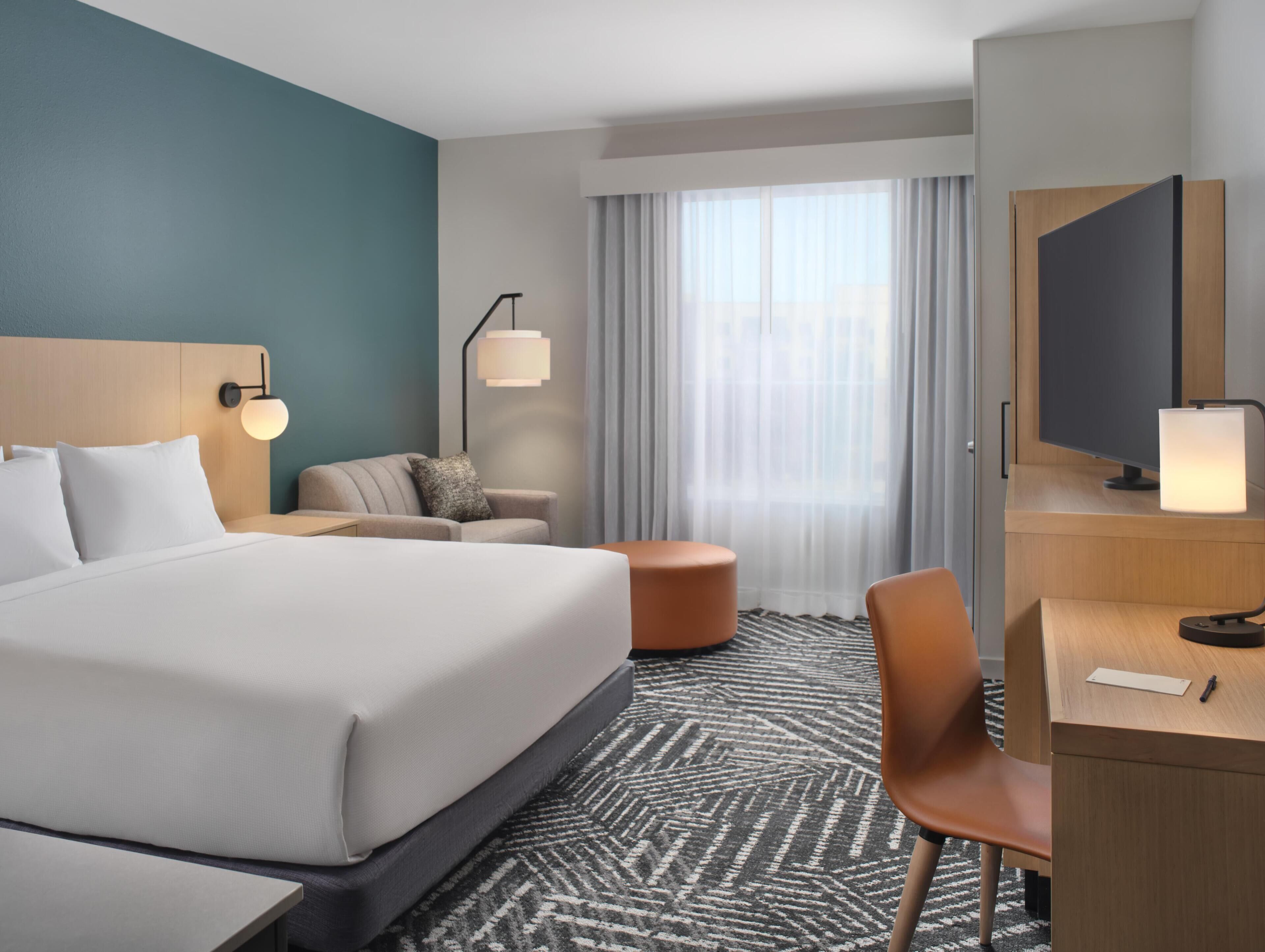 A hotel room with a large bed, a desk, a chair, a sofa, a TV, and a window with curtains.