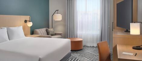 A hotel room with a large bed, a desk, a chair, a sofa, a TV, and a window with curtains.