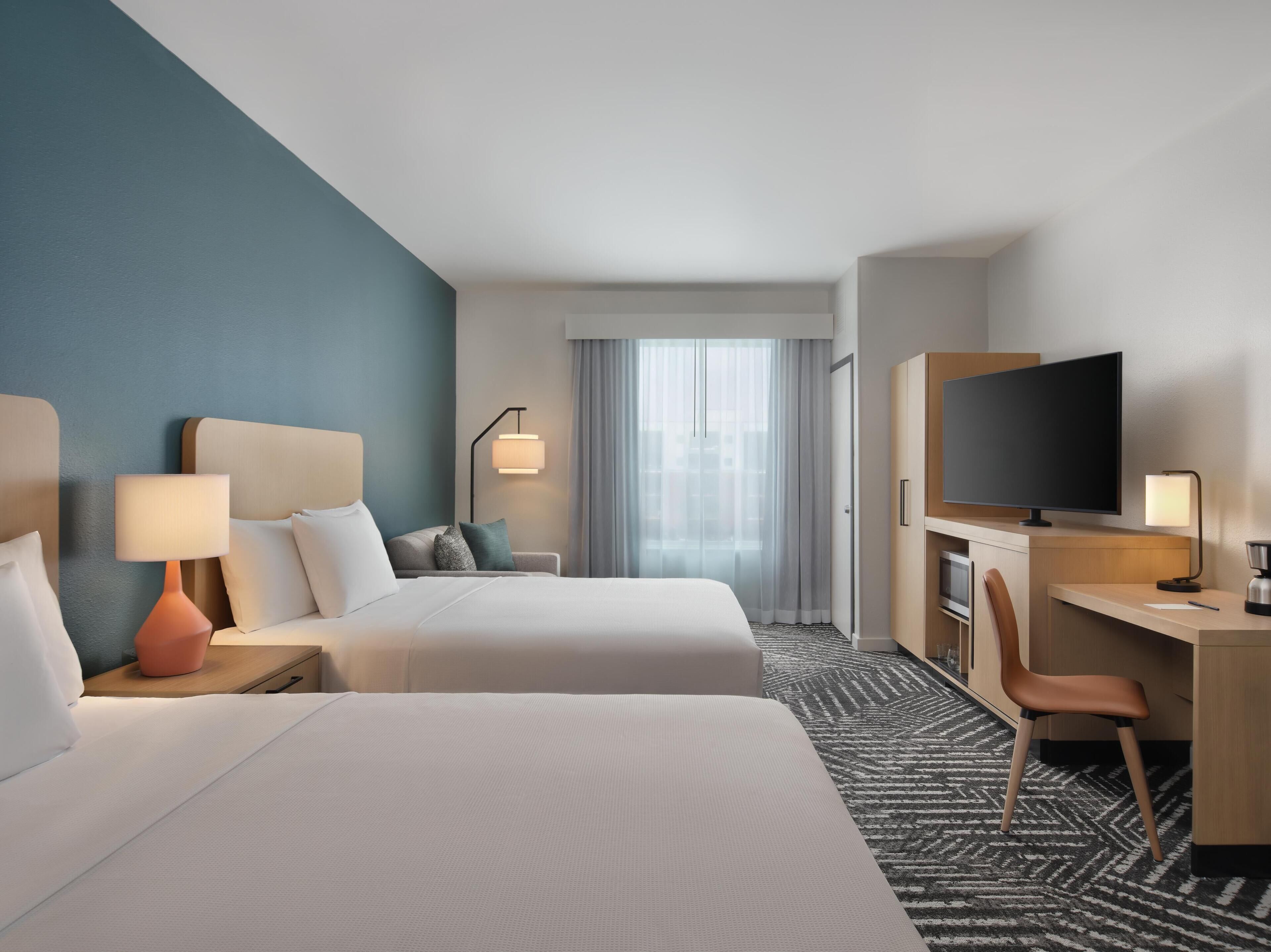 A hotel room with two beds, a desk with a TV, a chair, a desk lamp, and a window with curtains.