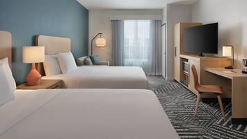 A hotel room with two beds, a desk with a TV, a chair, a desk lamp, and a window with curtains.