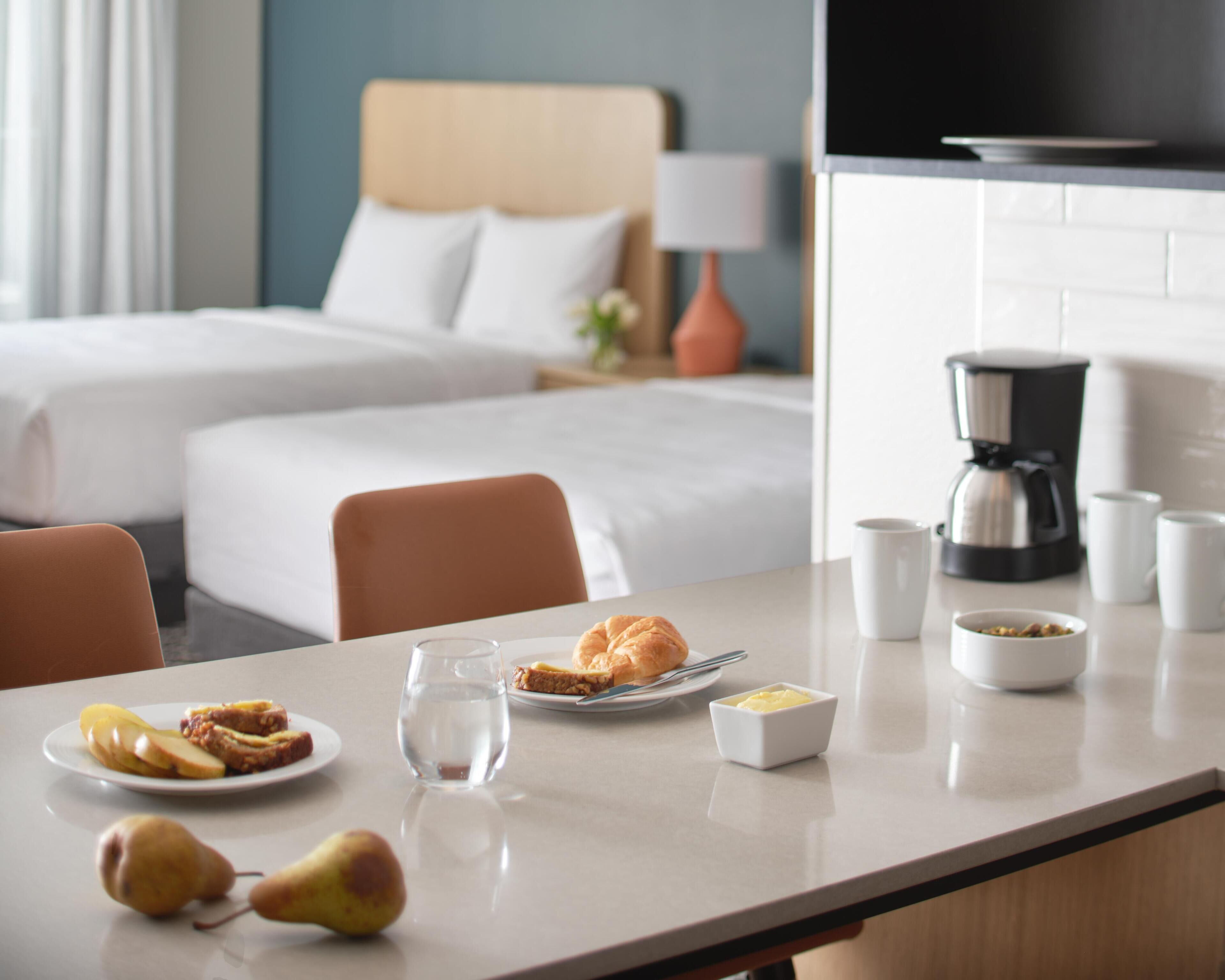 A hotel room with two beds, a dining table with breakfast items, a coffee maker, and a view of the city.