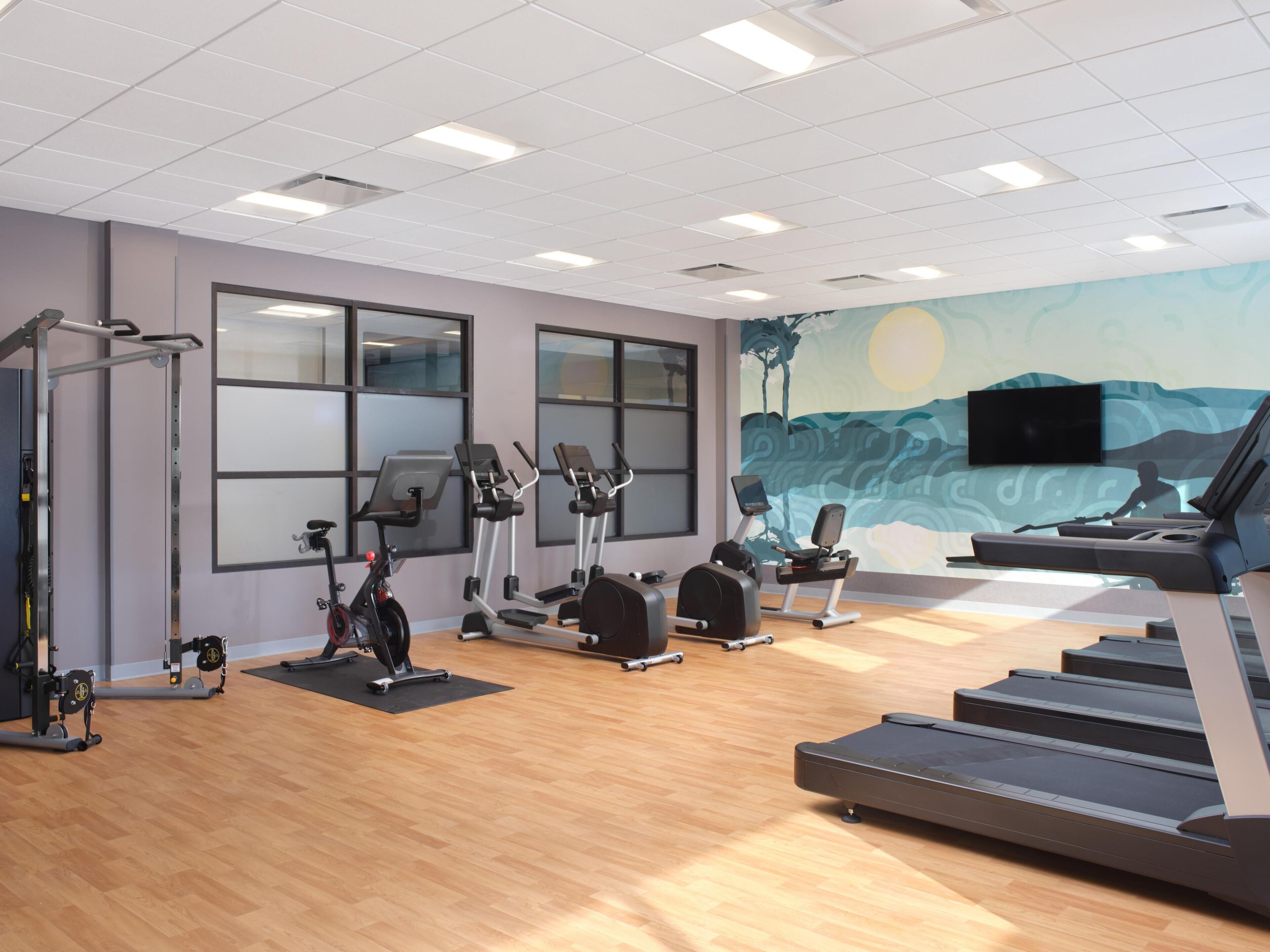 Ein Fitnessraum mit Trainingsgeräten, großen Fenstern und einem Wandgemälde.
